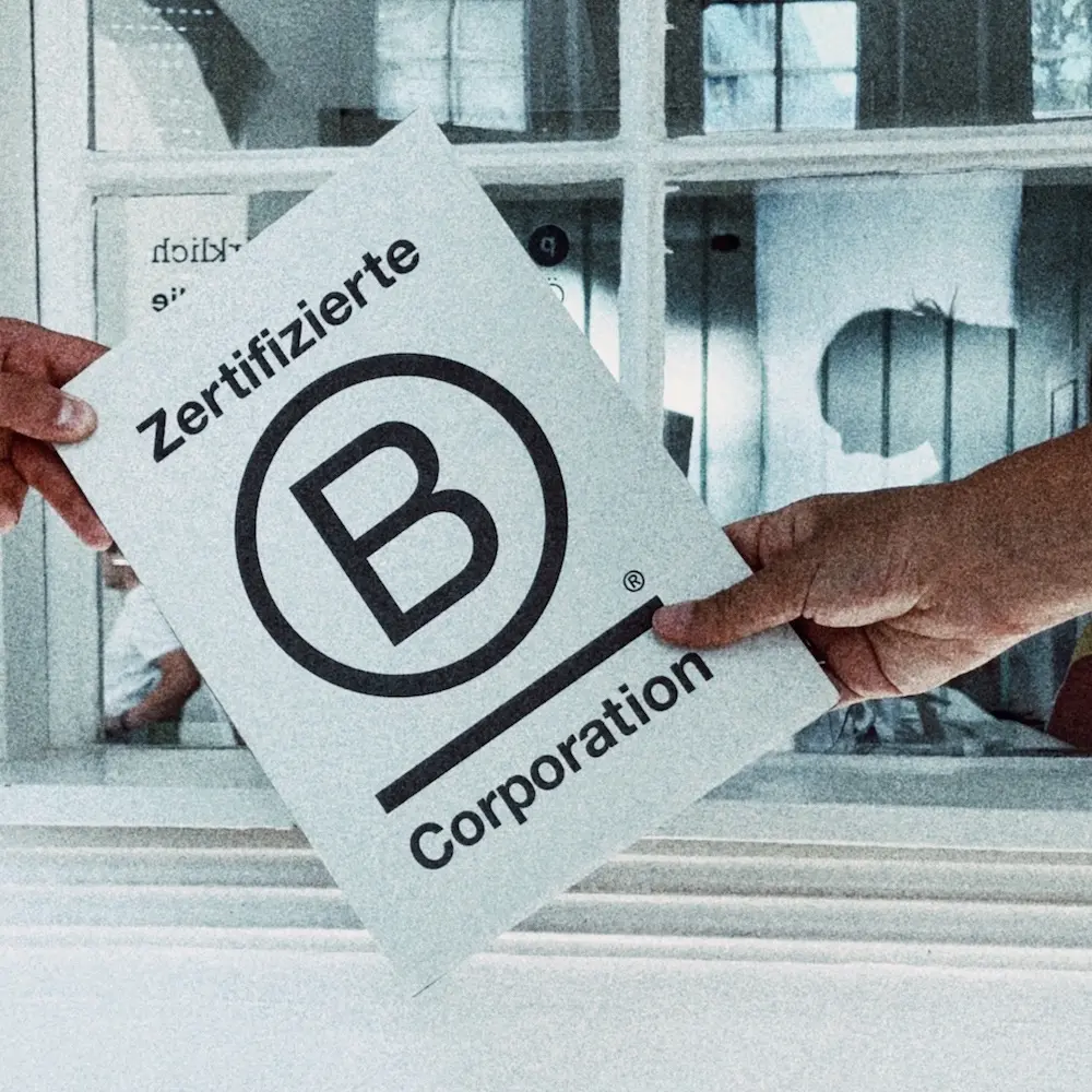 Zwei Hände halten ein kleines Poster mit dem B Corp-Logo vor einem Büro-Innenraum-Fenster.
