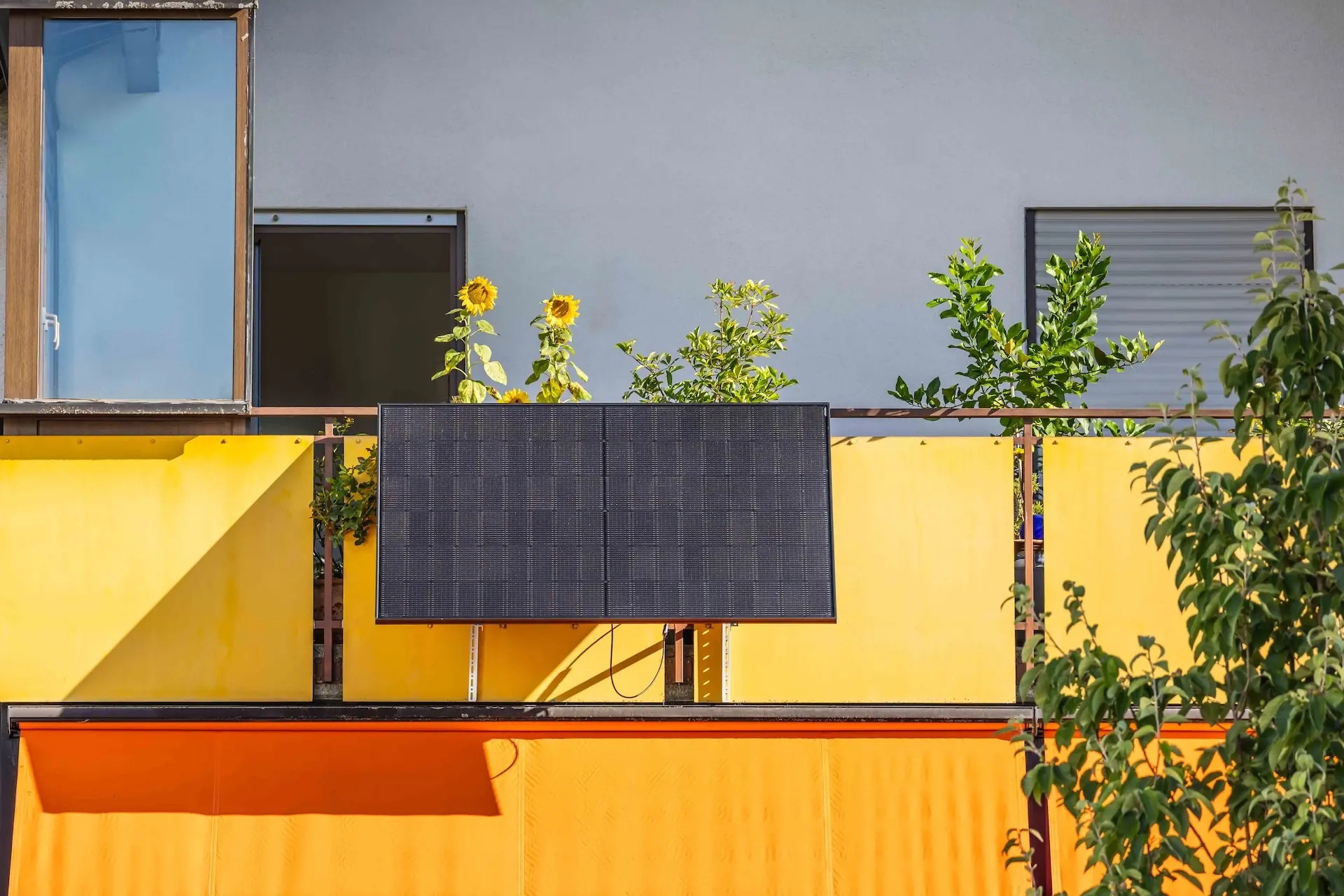 Ein schwarzes Solarpanel-Array, das an einem gelben Balkon mit einer orangefarbenen Markise und Sonnenblumen montiert ist.