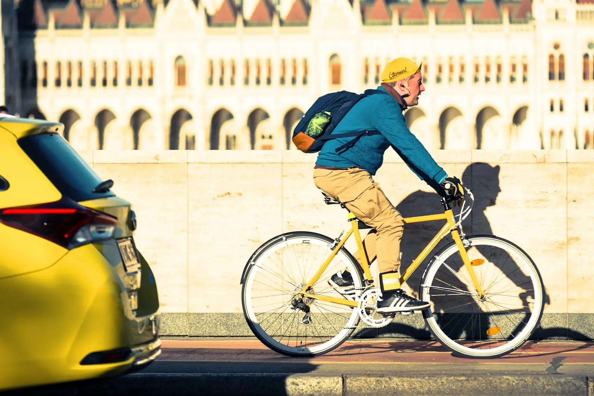 Ein Mann auf einem gelben Fahrrad fährt an einem gelben Auto vorbei, im Hintergrund steht ein prunkvolles Gebäude.