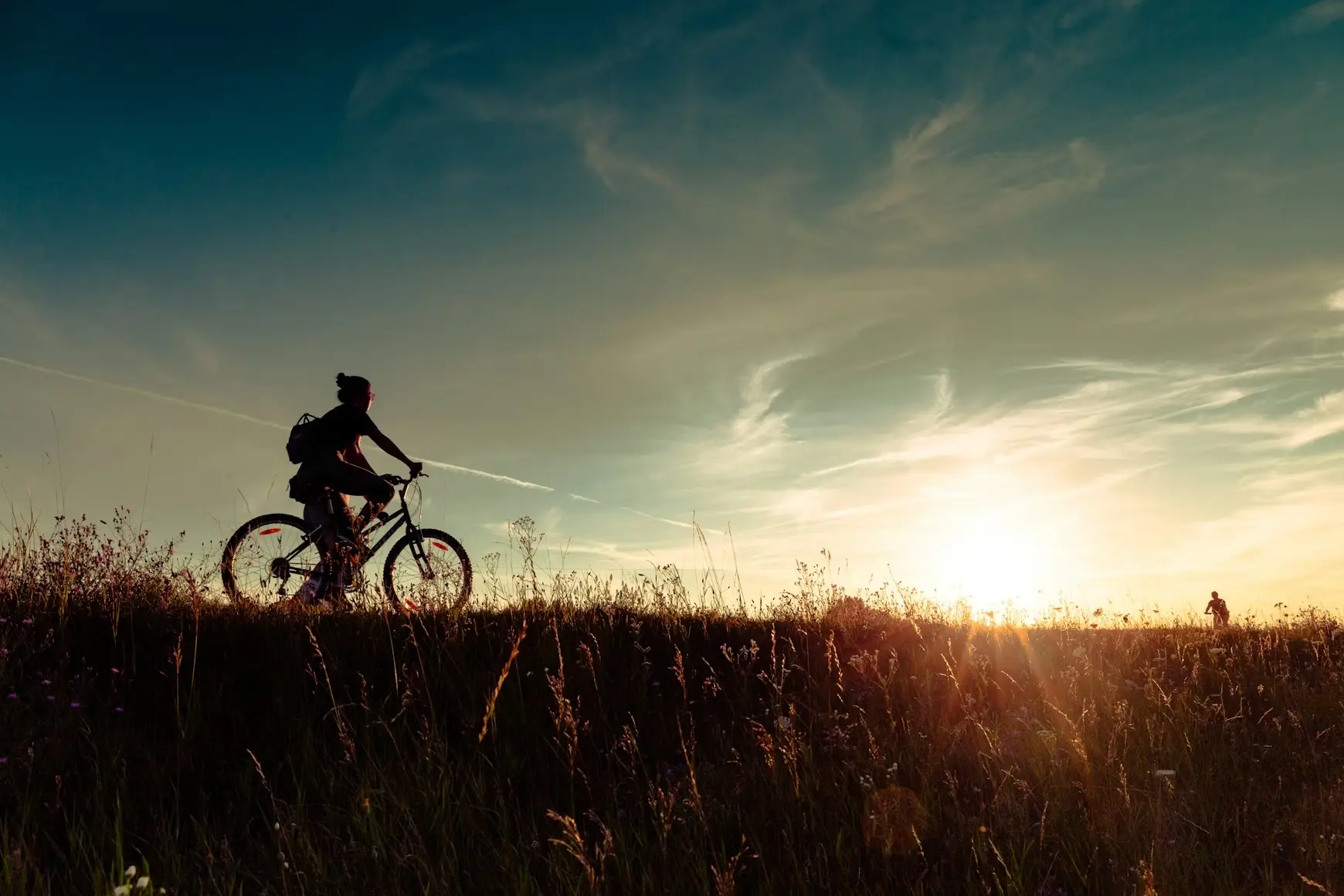 Silhouette einer Person, die bei Sonnenuntergang mit einem Fahrrad über ein Feld fährt.
