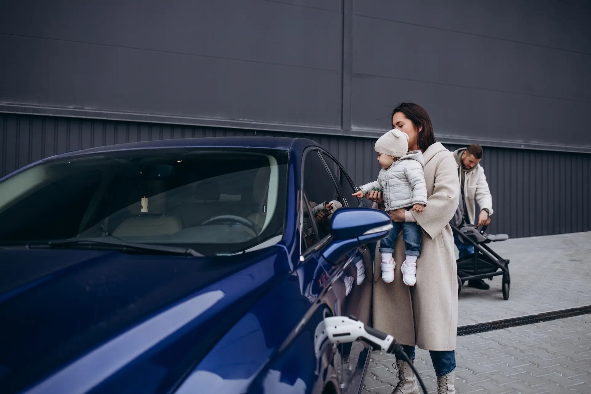 Eine Frau lädt mit einem Ladekabel das E-Auto, während der Mann das Kind ins Auto trägt