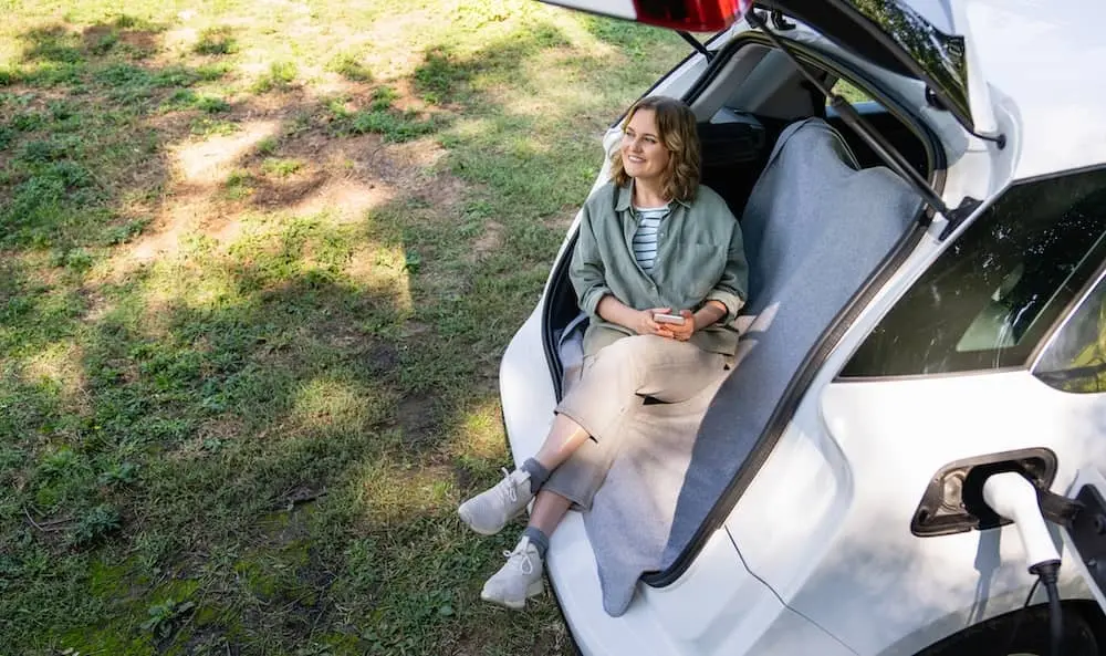 Eine Frau sitzt im offenen Kofferraum eines ladenden Elektroautos, das auf Gras geparkt ist.