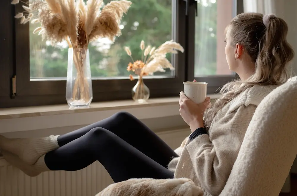 Eine Frau entspannt in einem gemütlichen Sessel, schaut aus dem Fenster und hält eine Tasse, während dekoratives Pampasgras auf der Fensterbank steht.