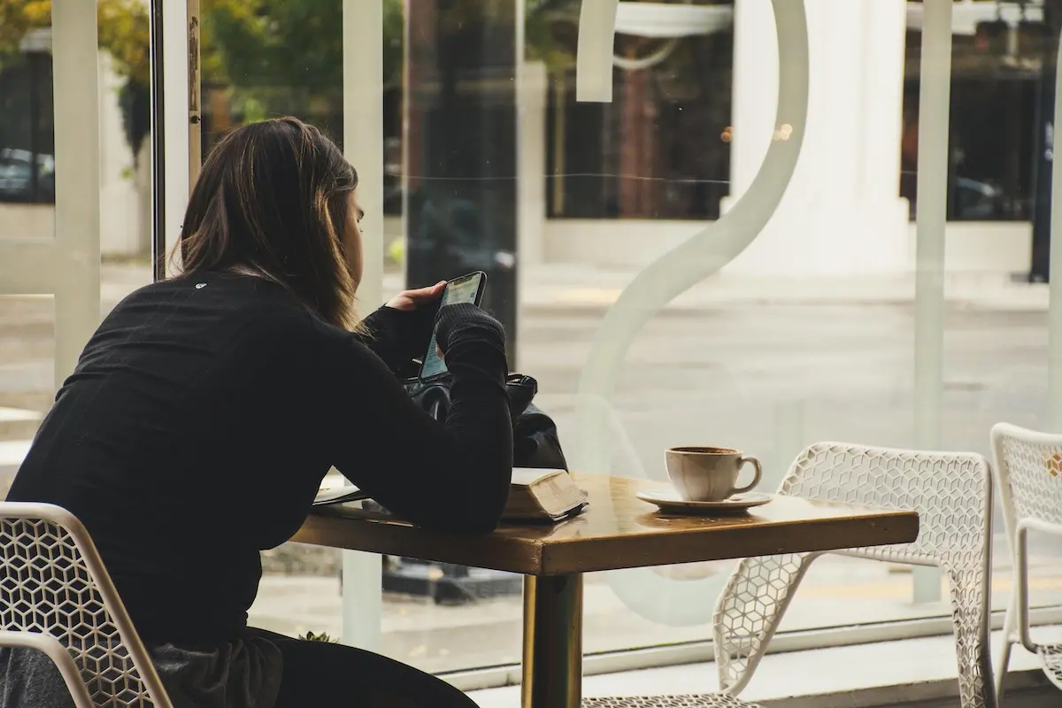 Eine junge Frau sitzt in einem Cafe am Tisch und nutzt ihr Handy. Vor ihr liegt ein augeschlagenes Buch.