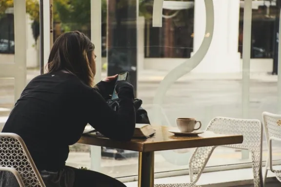 Eine junge Frau sitzt an einem Tisch und liest etwas auf dem Smartphone. Auf dem Tisch liegt eine aufgeschlagenes Buch und eine verschüttete Kaffeetasse.