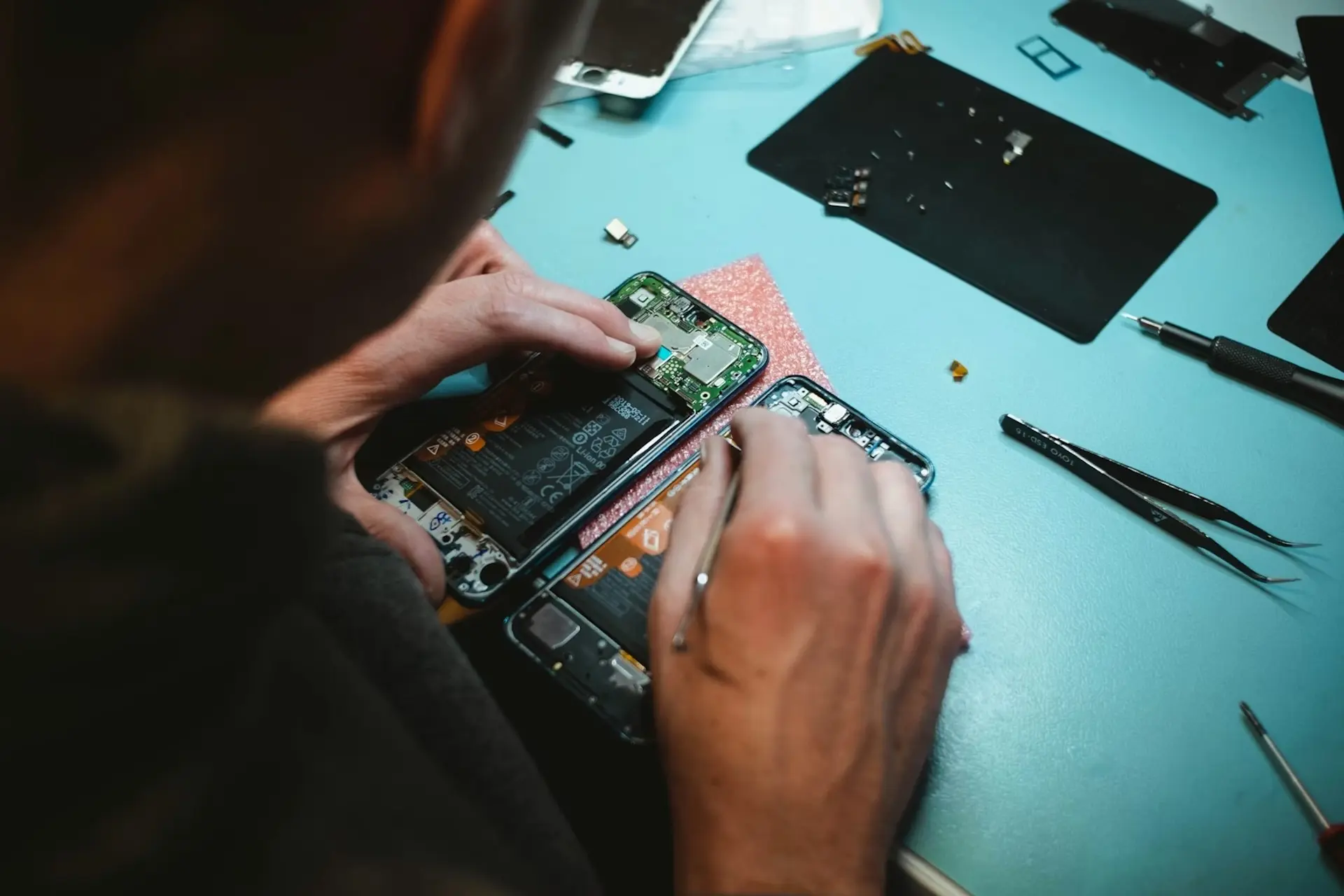 Hände, die zerlegte Smartphones auf einer blauen Werkbank mit verschiedenen Werkzeugen reparieren.
