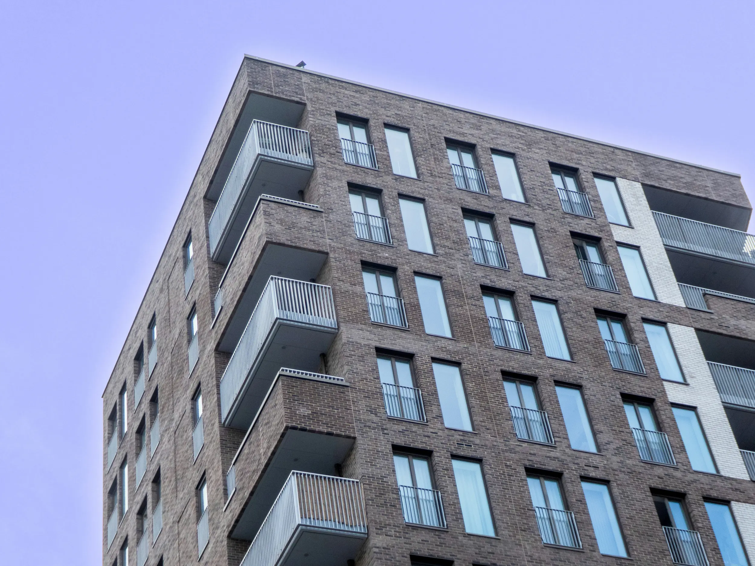 Oberer Teil eines modernen Mehrfamilienhauses mit Backsteinfassade, mehreren Balkonen mit Metallgeländern und vielen Fenstern. Der Himmel im Hintergrund ist klar und violett gefärbt.