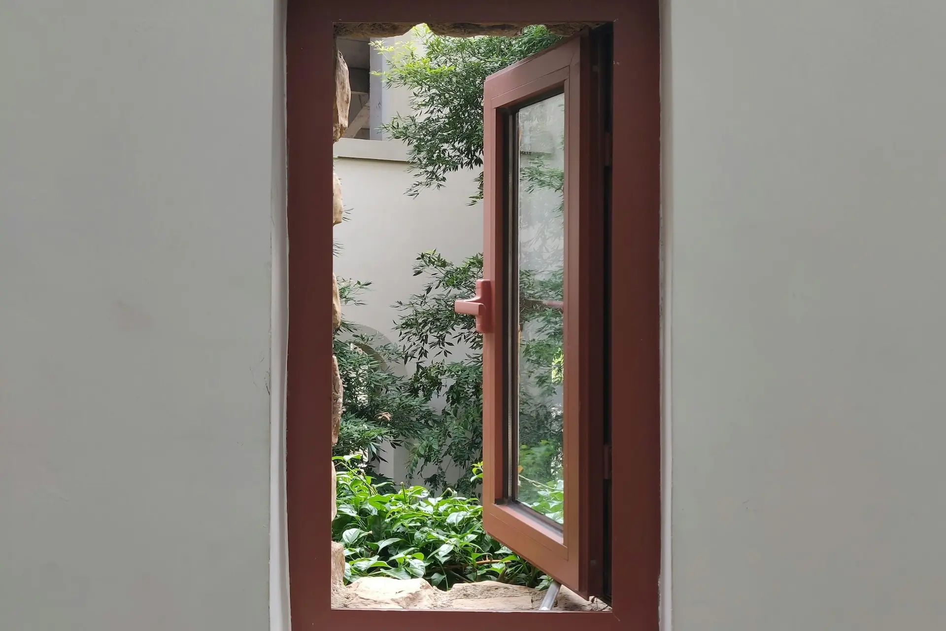Ein Fenster mit braunem Rahmen steht offen zu einem südländischen Hof, der mit Pflanzen bewachsen ist