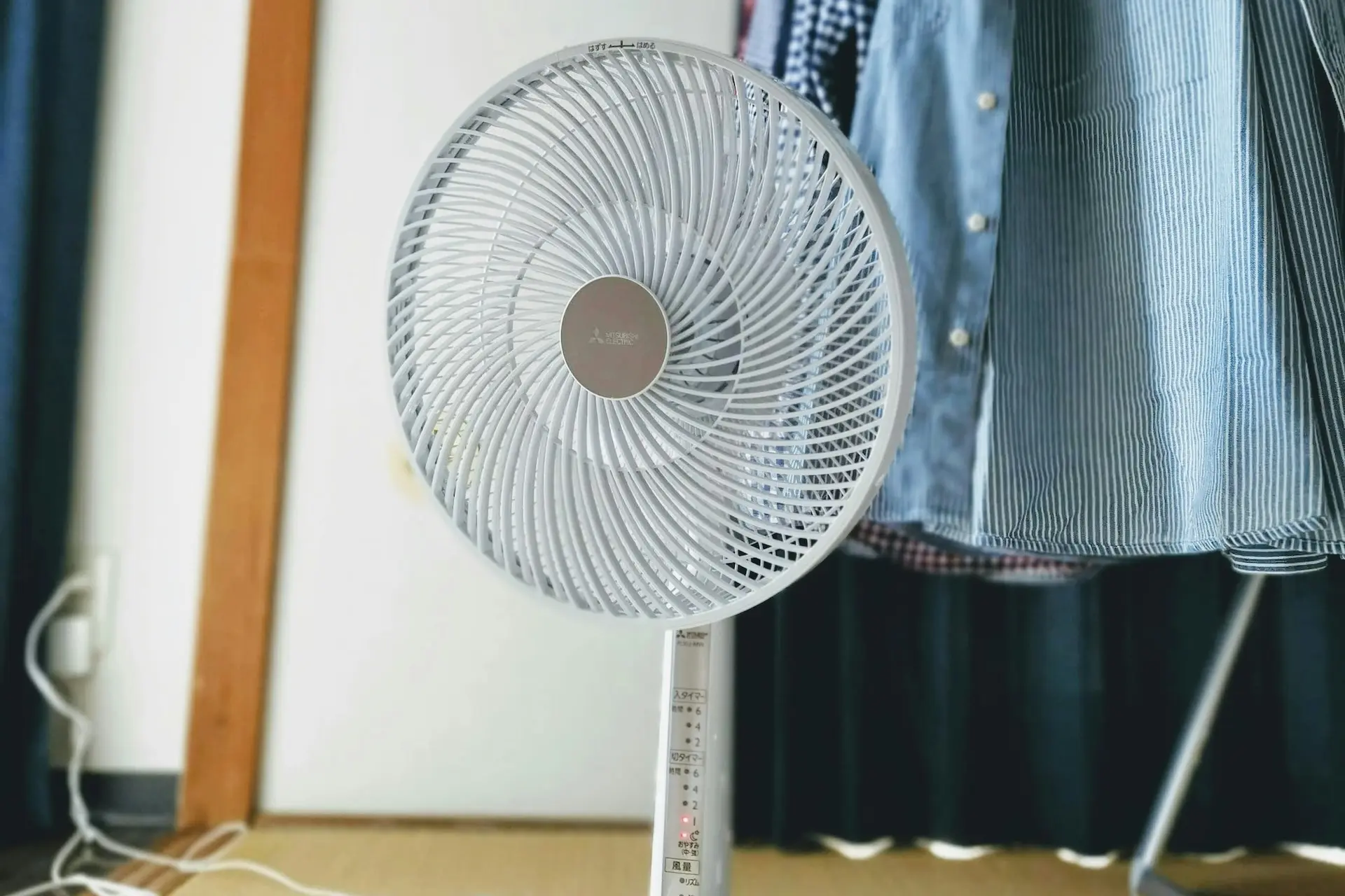 Ein weißer Standventilator steht in einem Raum vor einer Wand. Im Hintergrund sind Kleidungsstücke auf einem Kleiderständer zu sehen.