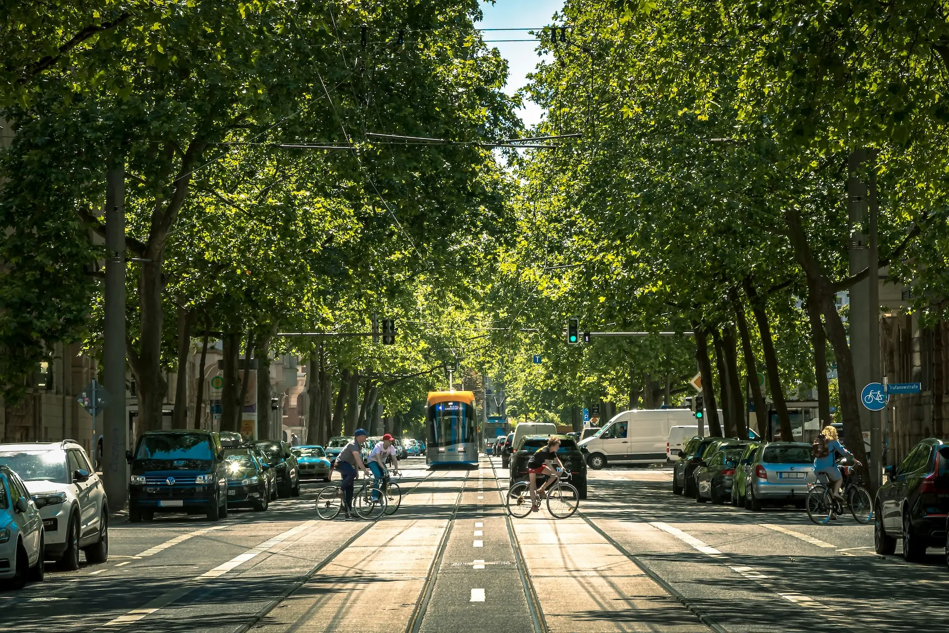 Eine sonnige Stadtstraße, gesäumt von üppig grünen Bäumen, mit einer Straßenbahn, Autos und Radfahrern auf der Straße.